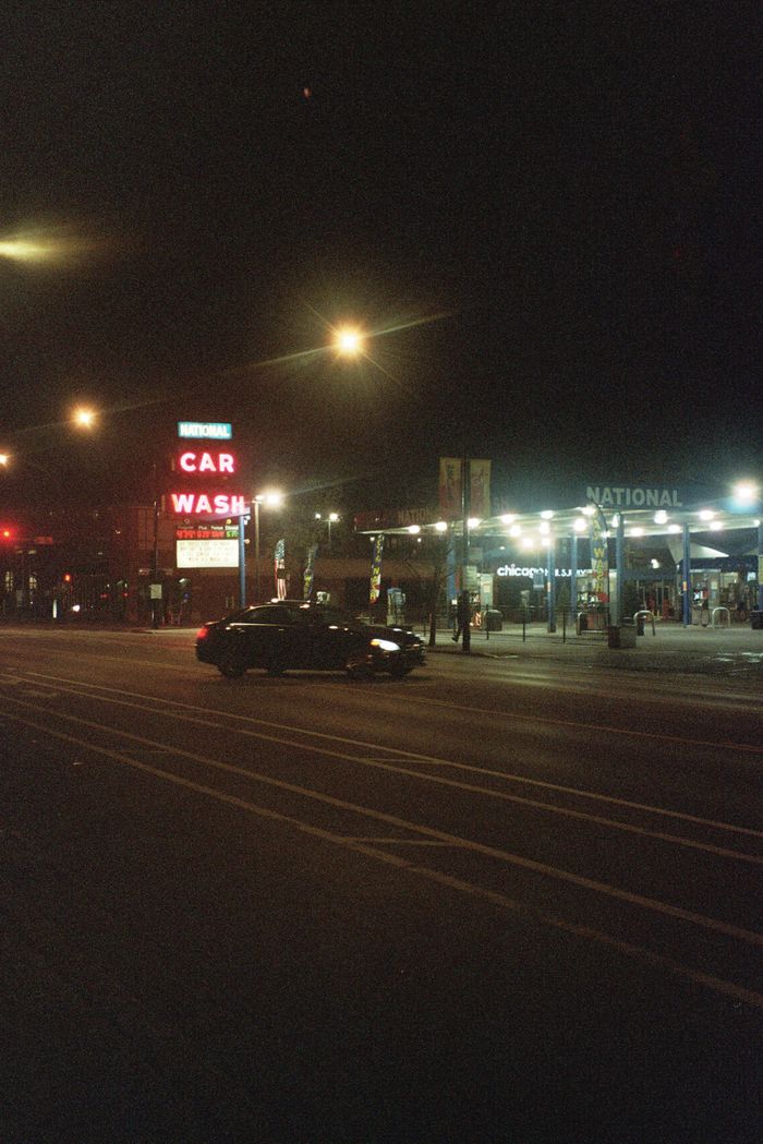 A black car passing by a gasoline station at night in Chicago, IL, illuminated by street lights.